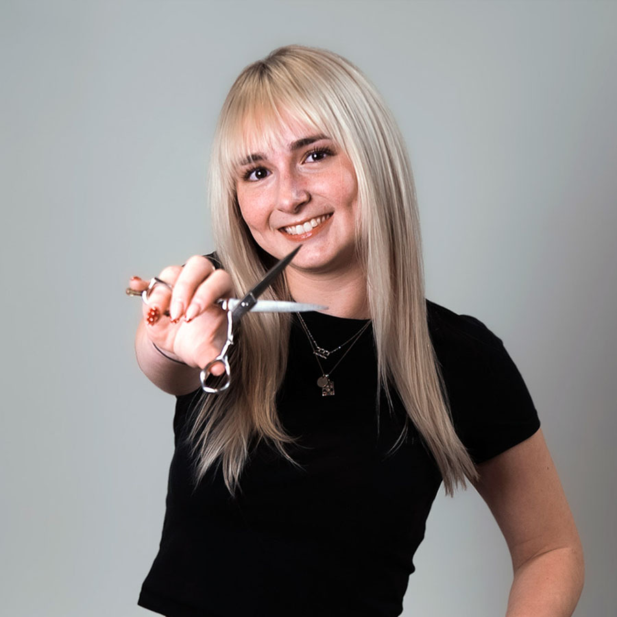 Smiling woman with long blonde hair extends a pair of scissors toward the camera, as if offering a haircut.