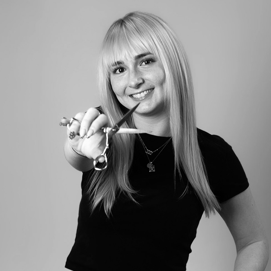 Smiling woman with long blonde hair wearing a black top, extending scissors toward the camera.