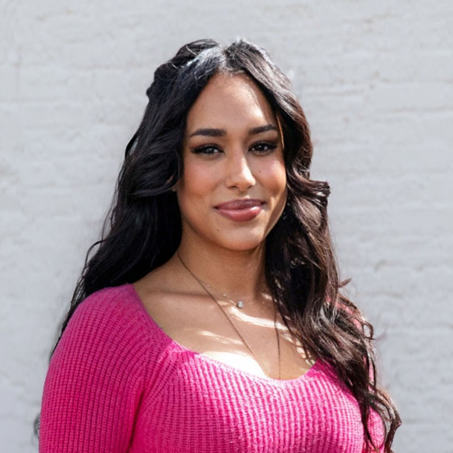 Portrait of a woman with long dark hair in a pink knit sweater, smiling outdoors.