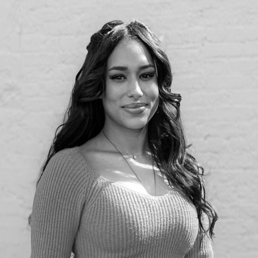 Portrait of a smiling woman with long dark hair wearing a knit sweater, standing outdoors against a brick wall.