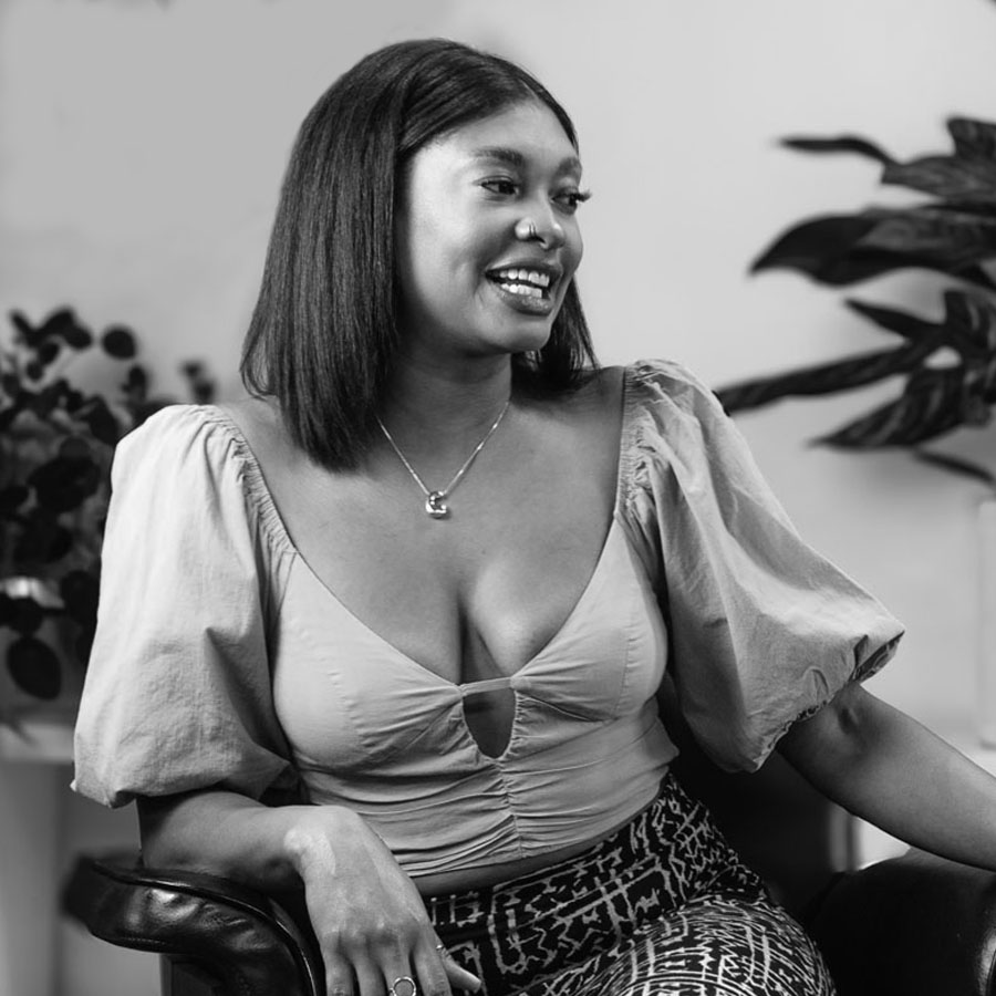 Woman smiling while seated in a chair, wearing a light top with puff sleeves and a patterned skirt, indoors with plants in the background.