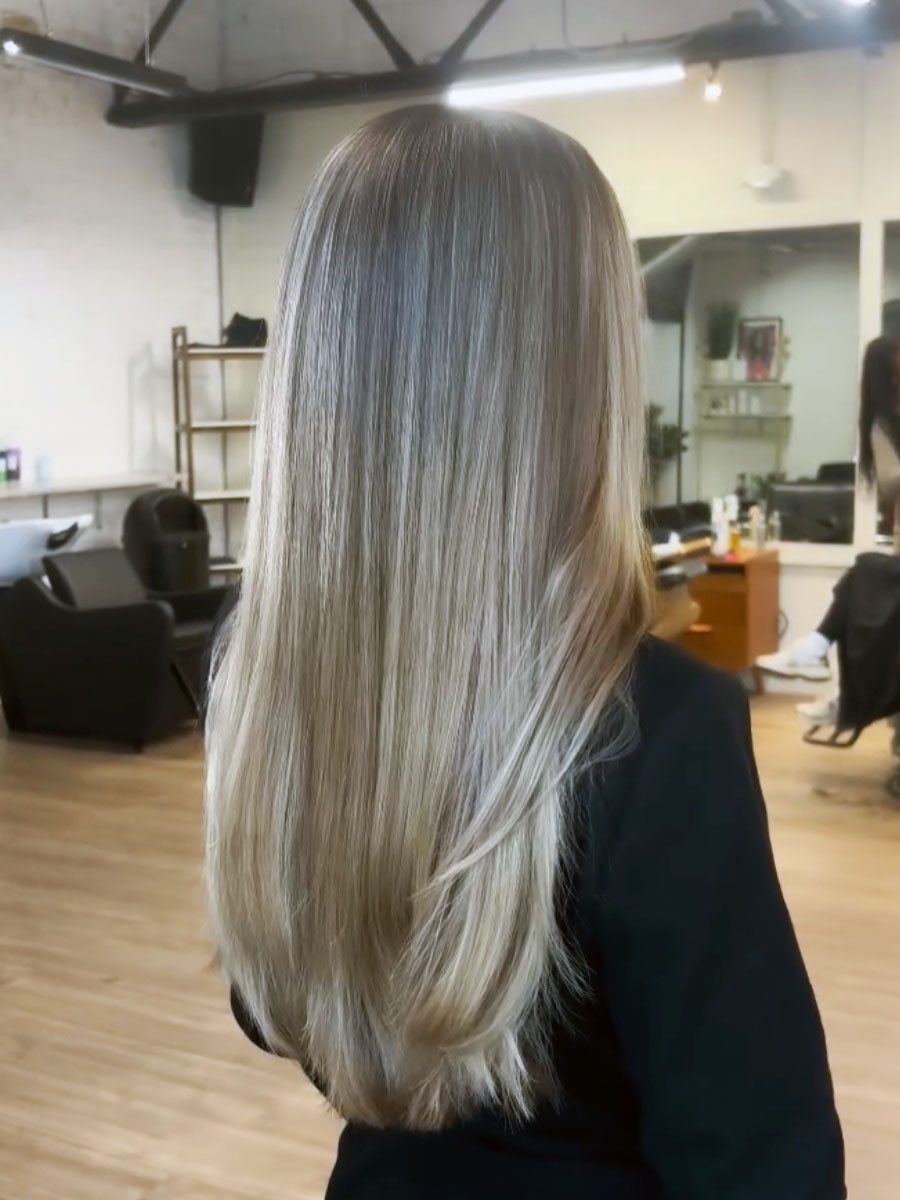 Back view of a woman with long straight blonde hair in a salon, featuring a cool-toned balayage and subtle darker roots.