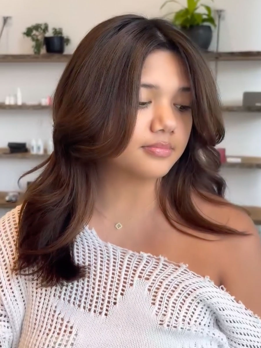 Portrait of a woman with shoulder-length brown hair looking down, wearing a white off‑the‑shoulder knitted top, in a bright room with shelves and plants in the background.
