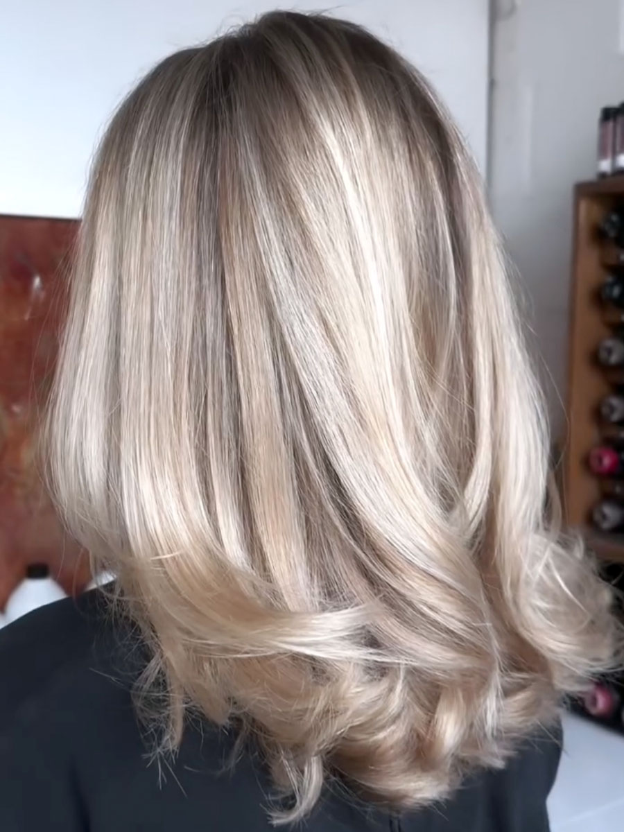 Back view of a shoulder-length blonde layered hairstyle with darker roots in a salon setting
