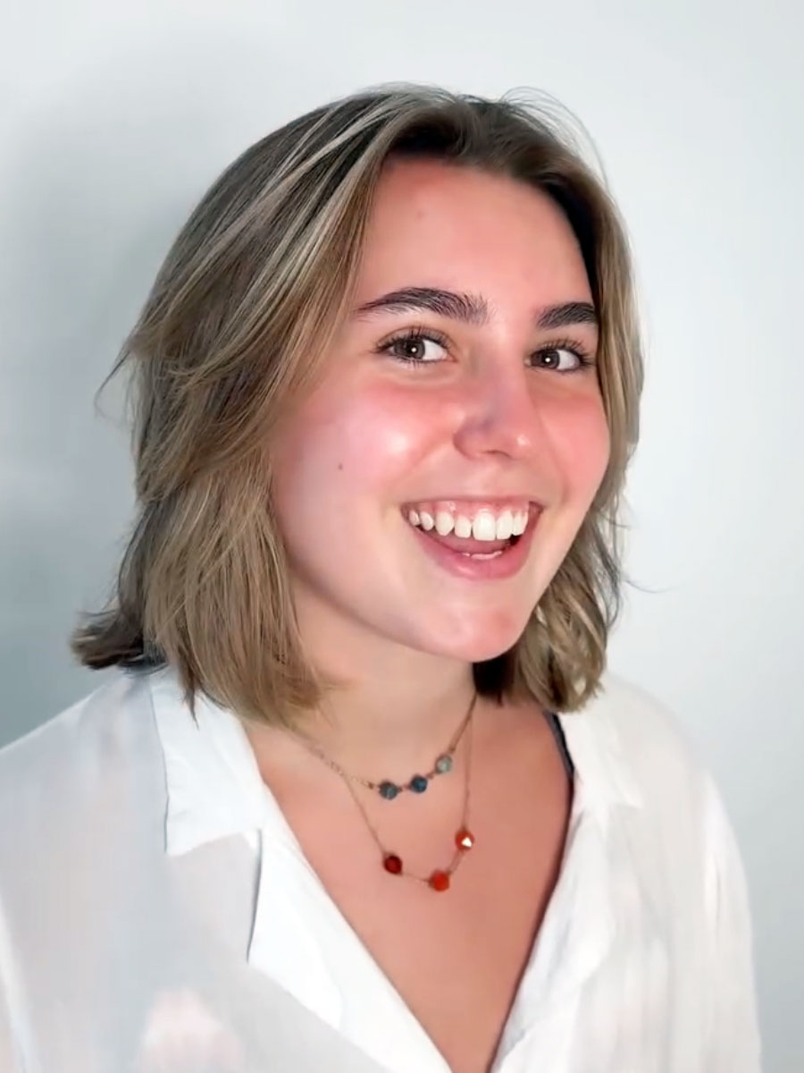Smiling young woman with short blonde hair wearing a white blouse and two beaded necklaces, facing the camera.