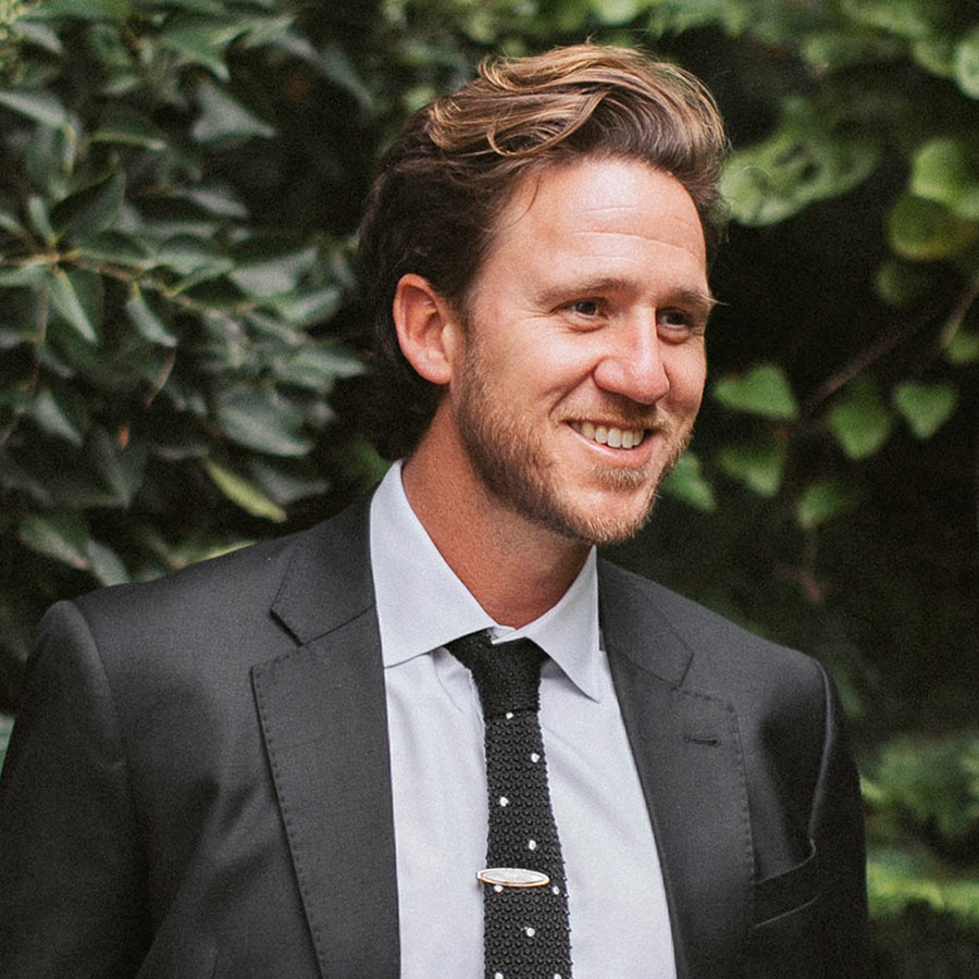 Smiling man in a dark suit and tie outdoors with greenery in the background, portrait-style image.