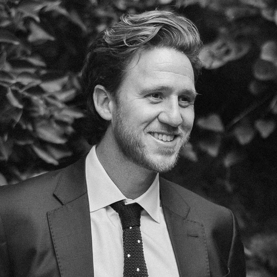 Smiling man in a dark suit and tie, outdoor portrait with leafy background.