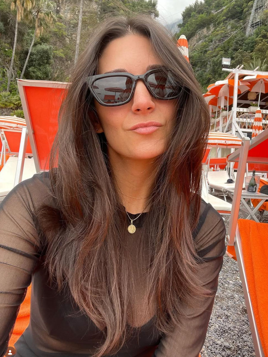 Woman with long dark hair wearing black sunglasses and a black sheer top at an outdoor beach setup with orange chairs behind her.