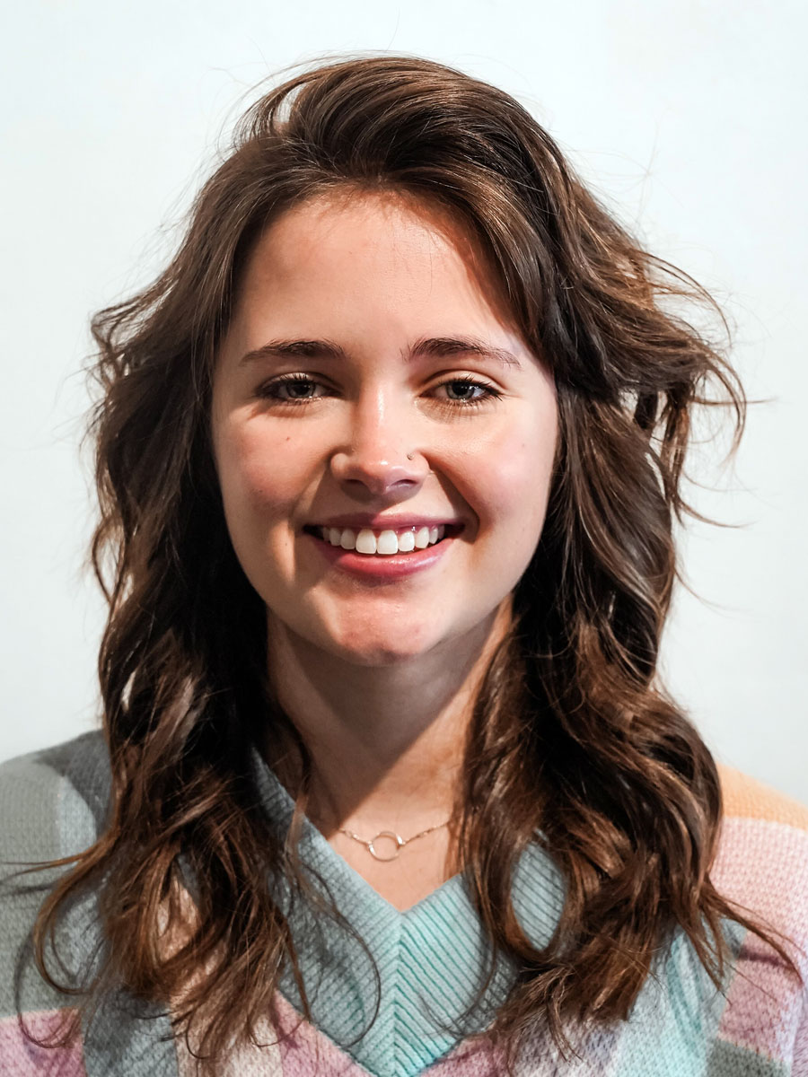 Portrait of a young woman with wavy brown hair smiling at the camera, wearing a pastel striped sweater.