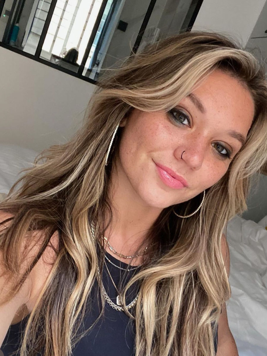 Selfie of a smiling woman with long wavy blonde hair, wearing large hoop earrings and a nose ring, in a bright bedroom.