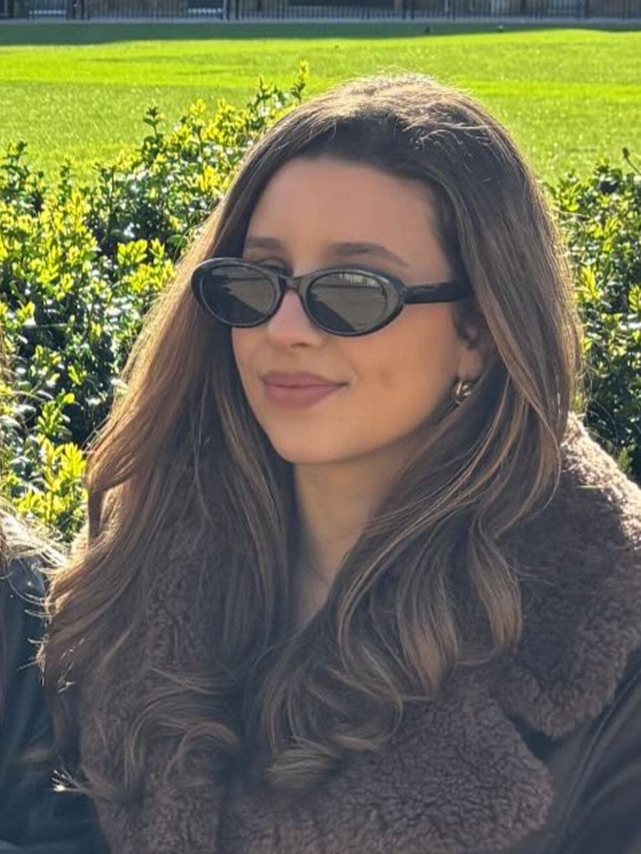Woman with long hair wearing oval sunglasses and a brown faux-shearling jacket, outdoors in a sunny park.