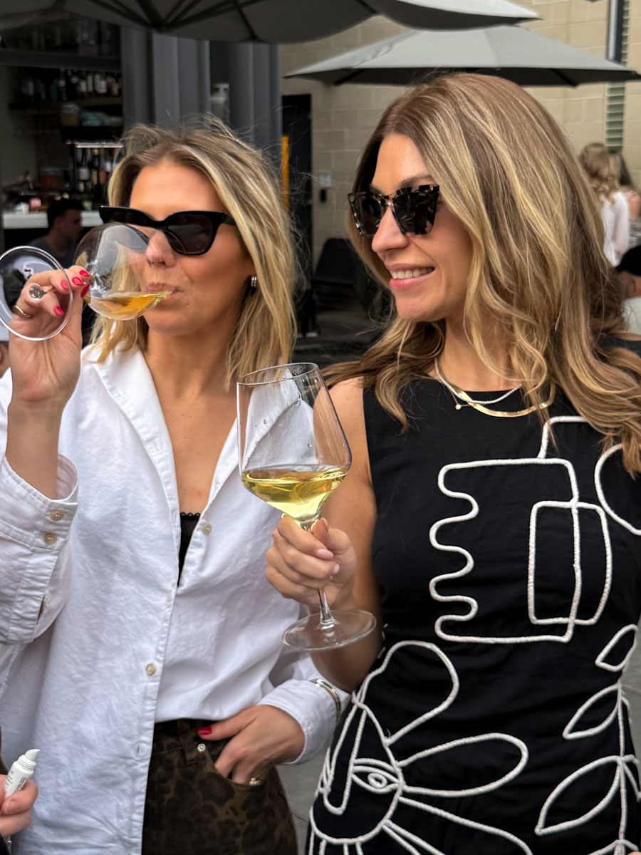 Two women wearing sunglasses toast with white wine at an outdoor bar under umbrellas, one in a white shirt and leopard-print bottom and the other in a black patterned dress.