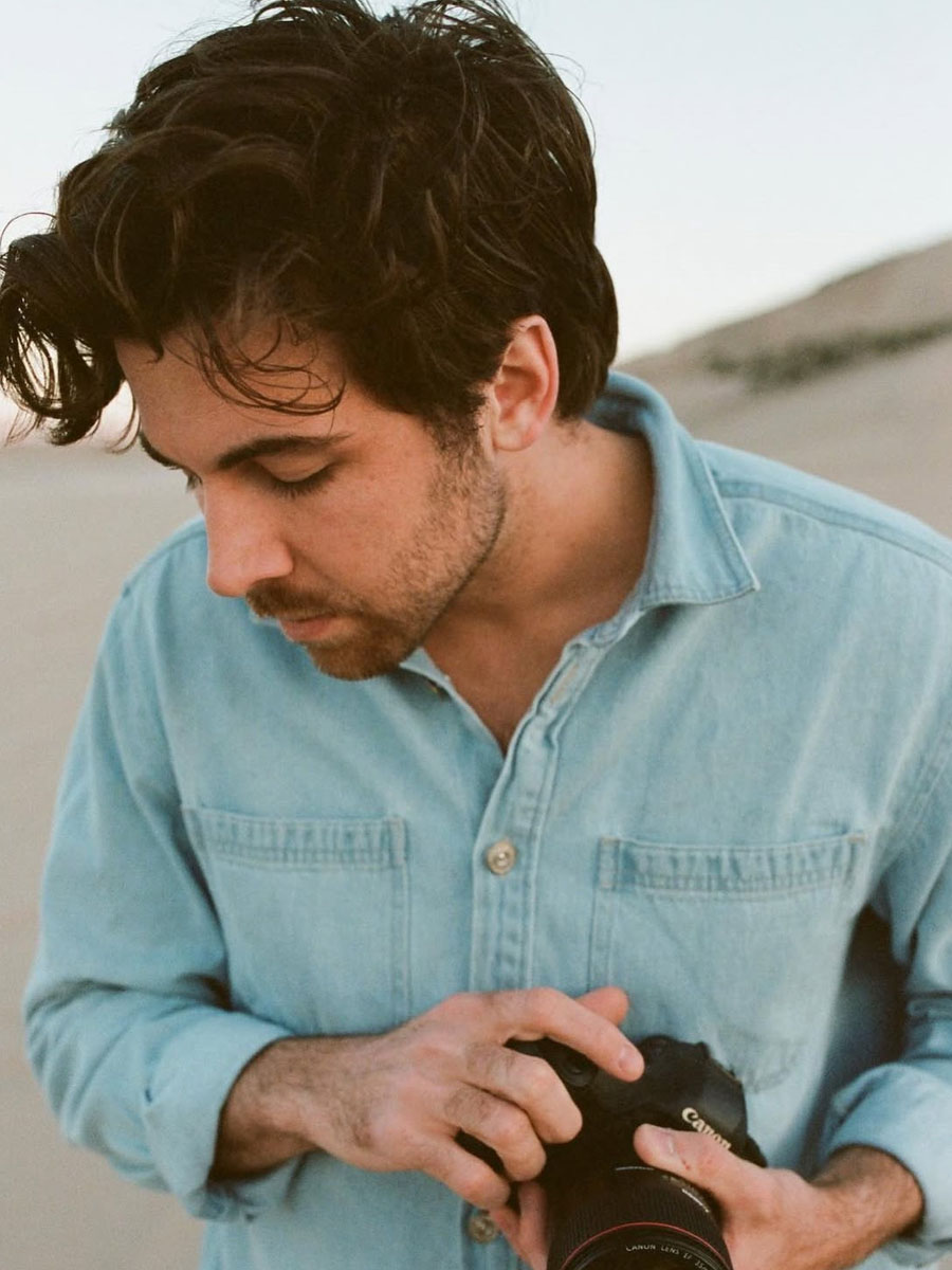 Man in a light blue denim shirt looking down while adjusting a black camera on a beach.