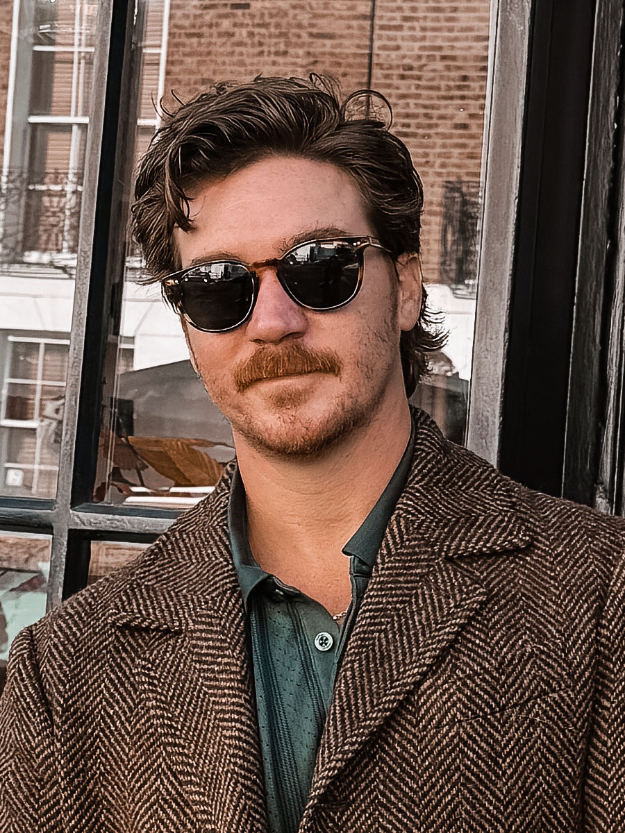 Man with brown hair and a mustache wearing round sunglasses and a brown herringbone coat outdoors by a brick building.