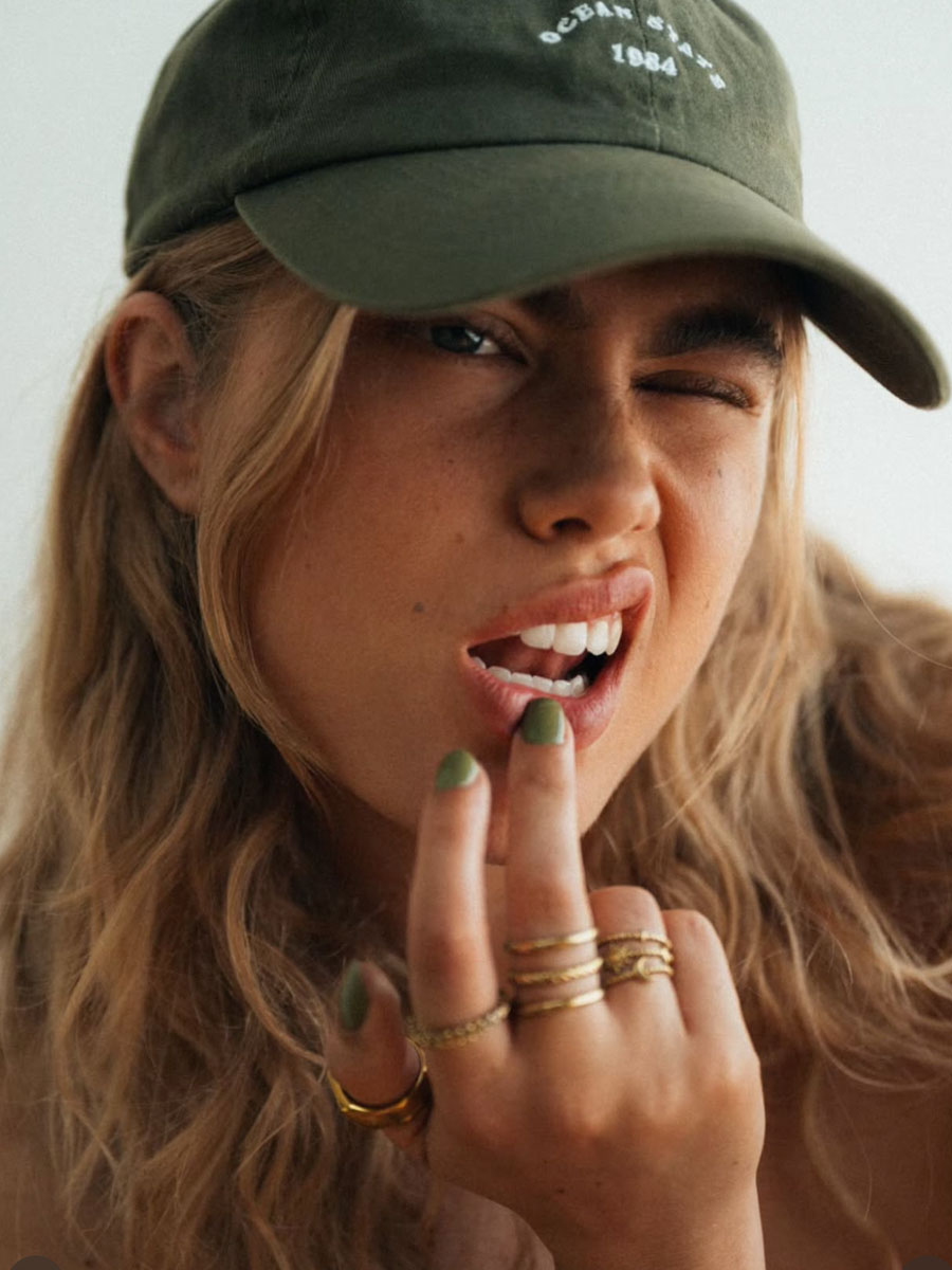 Young woman wearing a green cap winks at the camera, finger near her lips, showcasing green nail polish and multiple gold rings.