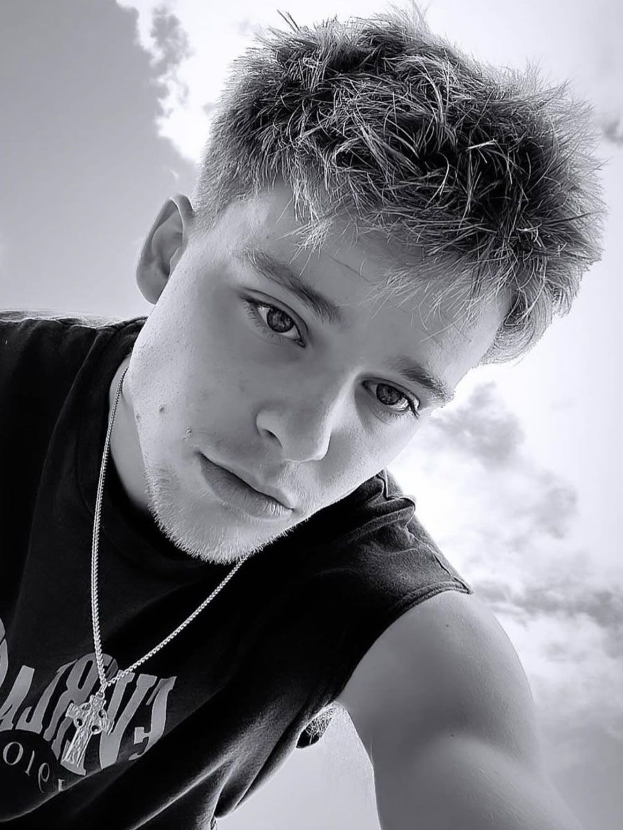 Young man with short spiky hair wearing a sleeveless black shirt and chain, taking a tilted selfie against a cloudy sky; black-and-white photo.