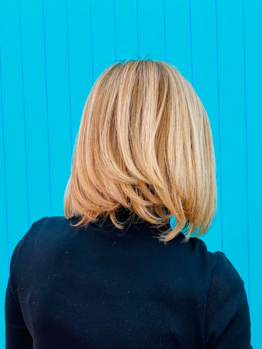 Back view of a person with shoulder-length blonde hair in a layered bob, wearing a dark top, against a bright blue fence/ wall.