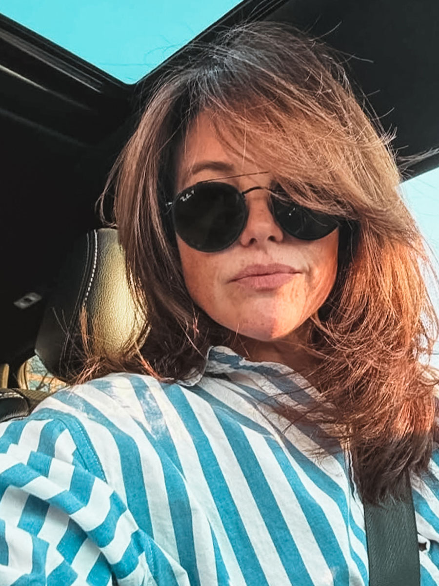 Woman wearing round black sunglasses in a car, striped blue-and-white shirt, seatbelt on, sunlight on her face.