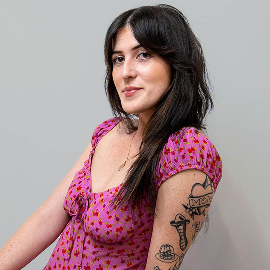 Portrait of a woman with dark hair wearing a pink floral dress, with tattoos visible on her forearm, posing against a gray background.