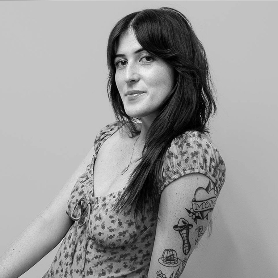 Portrait of a woman with long dark hair, wearing a floral dress, tattooed right arm visible with a 'MOM' heart tattoo and other designs.