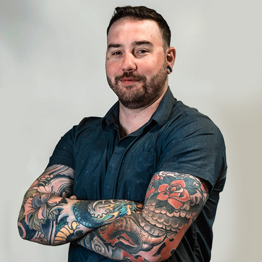Man with short dark hair and a beard, wearing a navy shirt with arms crossed, showing colorful full-arm tattoos.