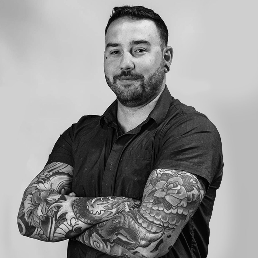 Man with short dark hair and beard, arms crossed, wearing a dark shirt, showing detailed full-arm tattoos in black and gray ink.