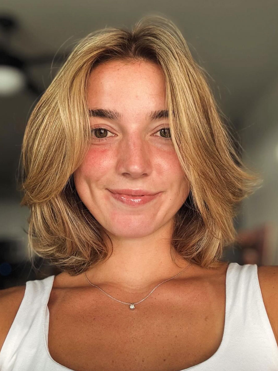 Smiling woman with shoulder-length blonde hair wearing a white top and a delicate necklace, indoors.