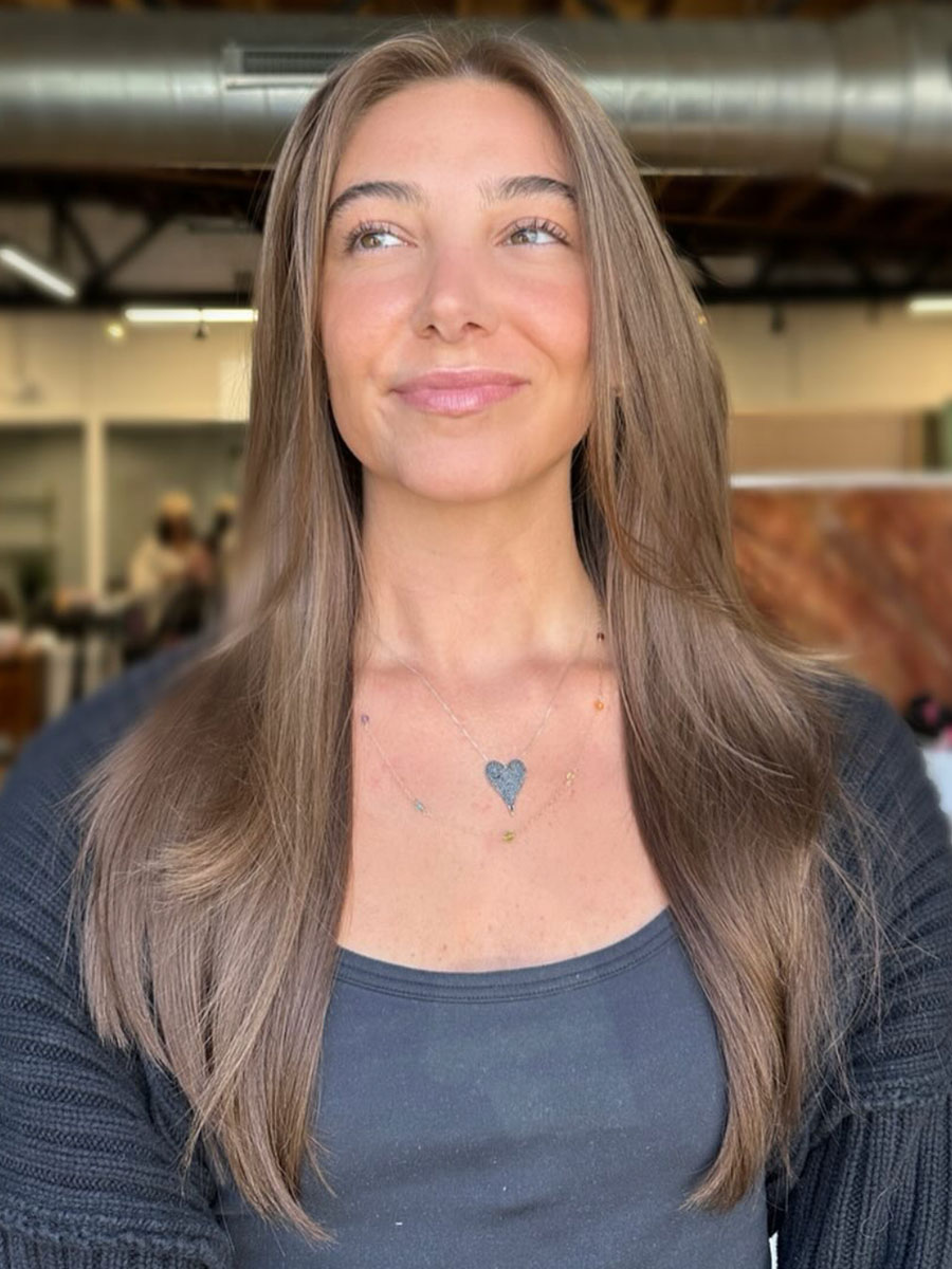 Young woman with long light brown hair, wearing a dark top and a heart pendant necklace, looking slightly upward in a bright indoor setting.