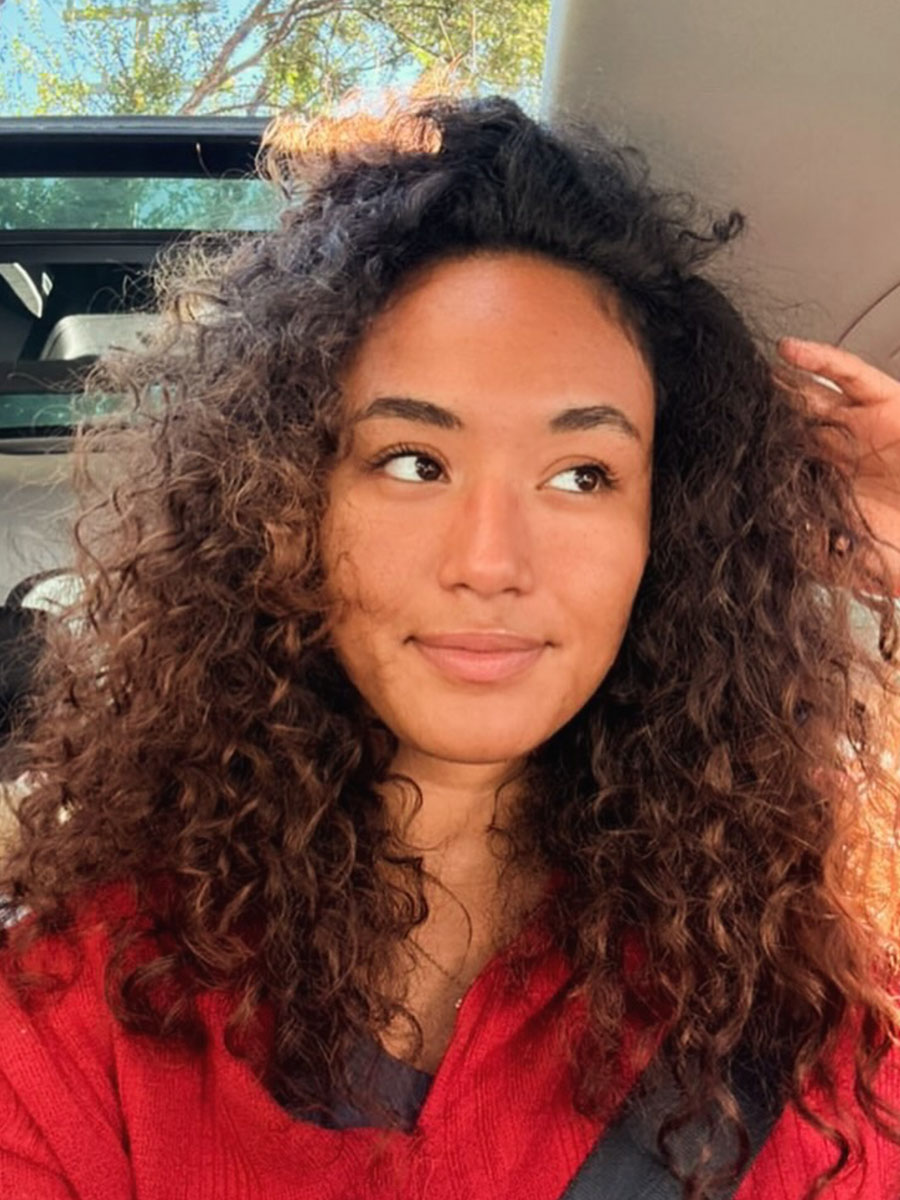 Young woman with curly hair in a car, wearing a red sweater and looking to the side with a gentle smile.