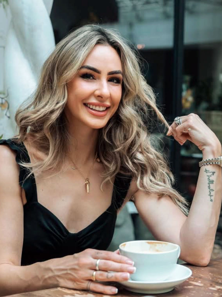 Smiling woman with wavy blonde hair sits at a cafe table with a cup of coffee in front of her.