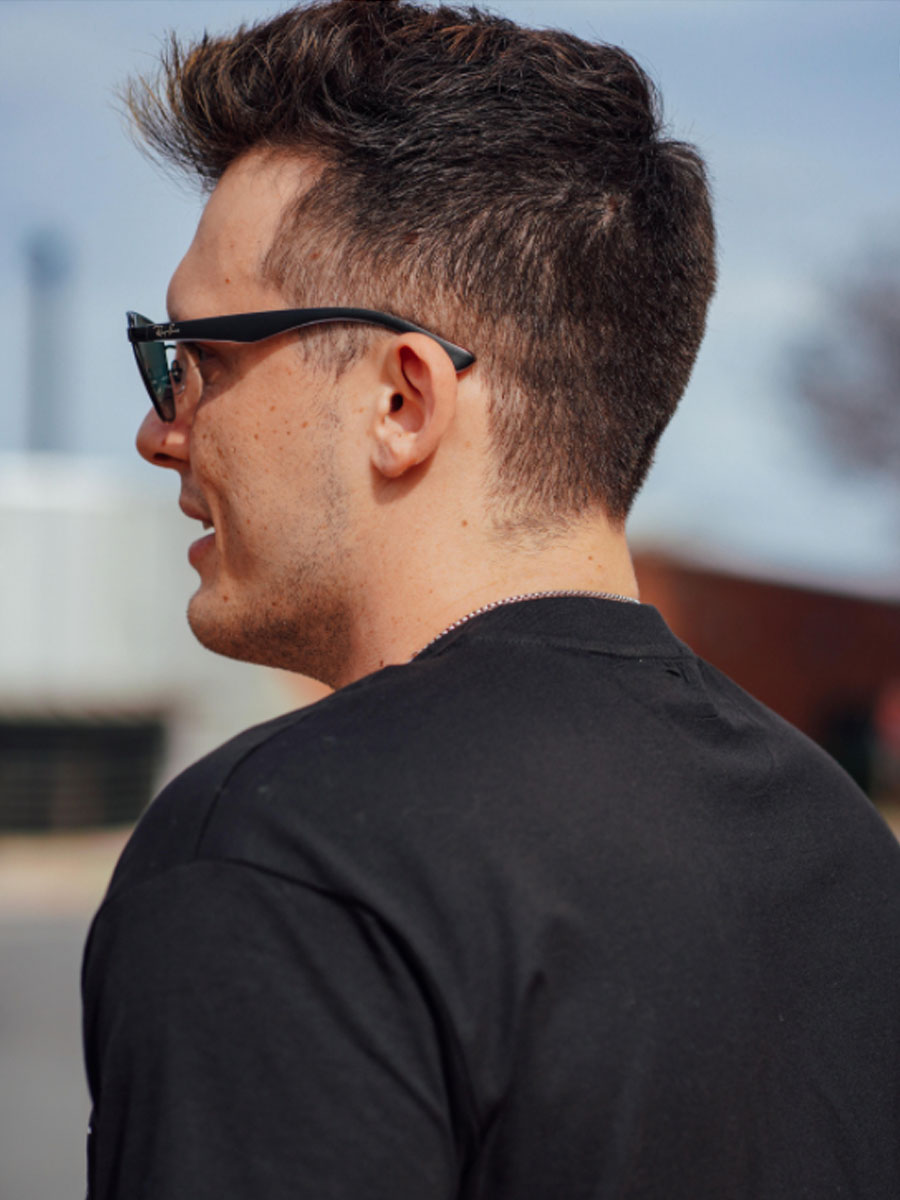 Profile of a man in black T-shirt wearing dark sunglasses, facing left outdoors.
