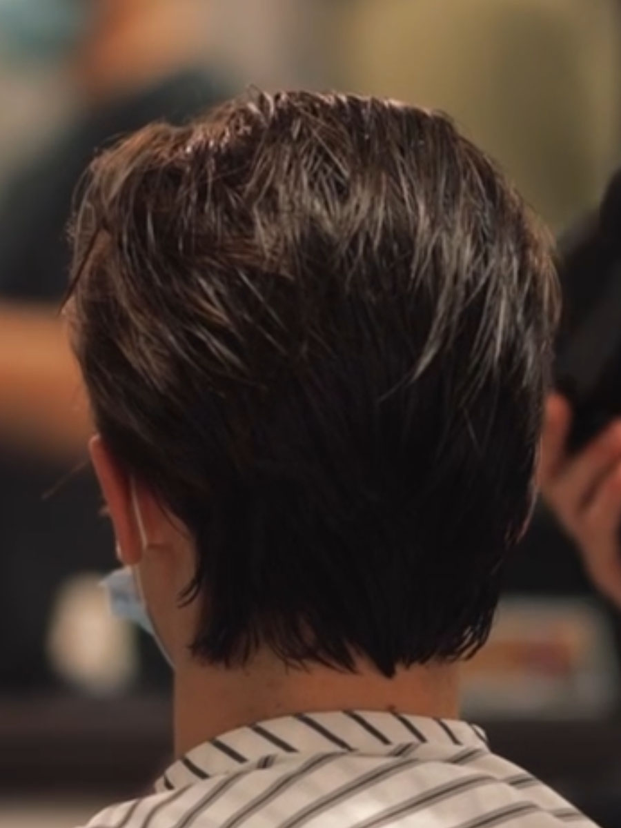 Back of a person’s head with short dark hair in a hair salon, wearing a striped collar shirt and a mask pulled under the chin.