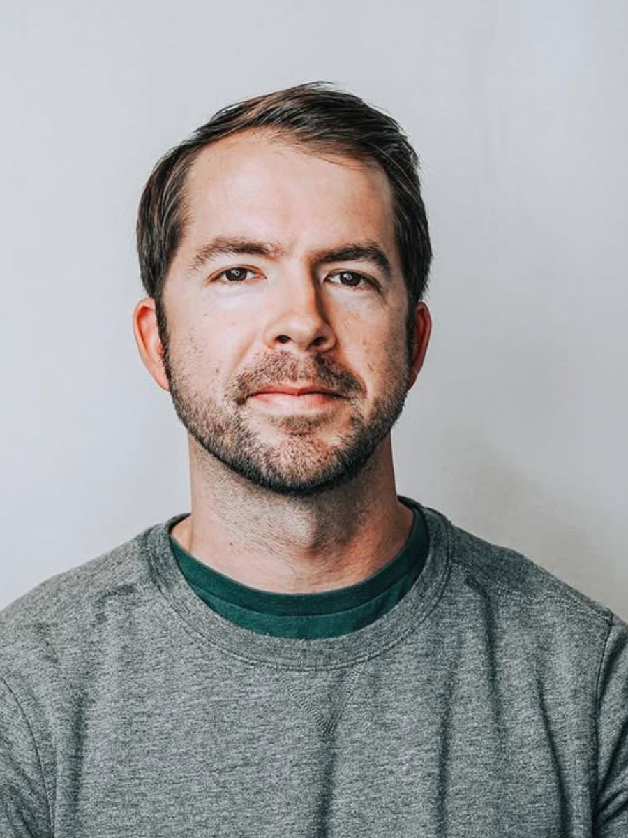 Portrait of a man with short dark hair and stubble, wearing a gray crew-neck sweater, looking at the camera against a light gray background.