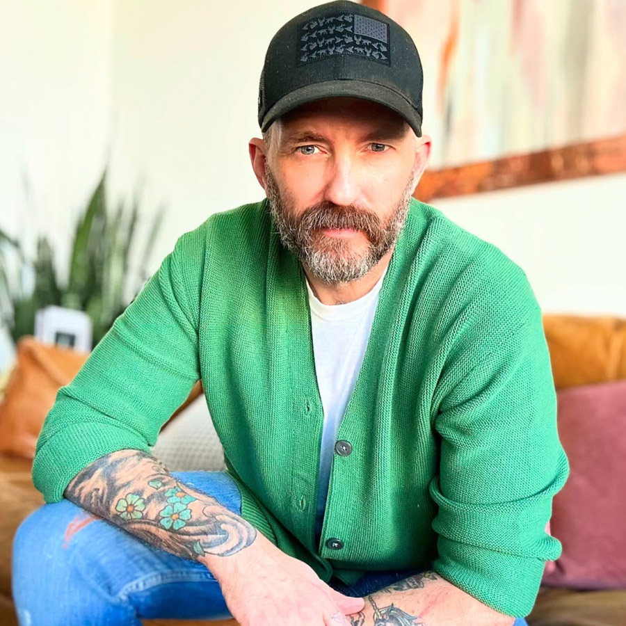 Man with a beard wearing a black cap and green cardigan, seated indoors with visible tattoos on his forearms.