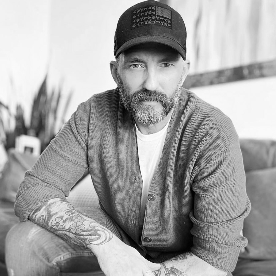 Portrait of a bearded man wearing a cap and cardigan, seated indoors, with tattoos visible on both forearms and a calm expression.