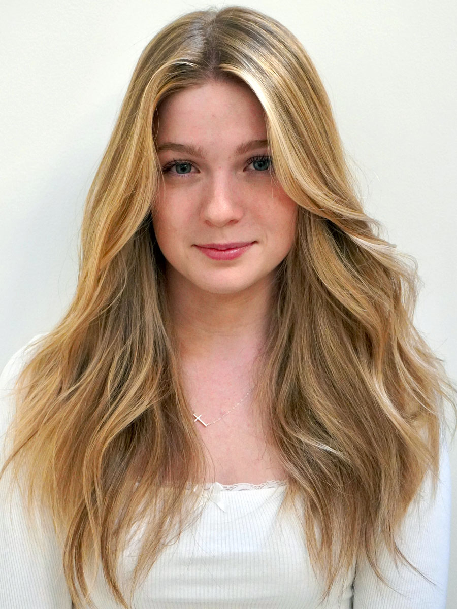 Portrait of a young woman with long wavy blonde hair, wearing a white top and a delicate necklace, facing the camera.