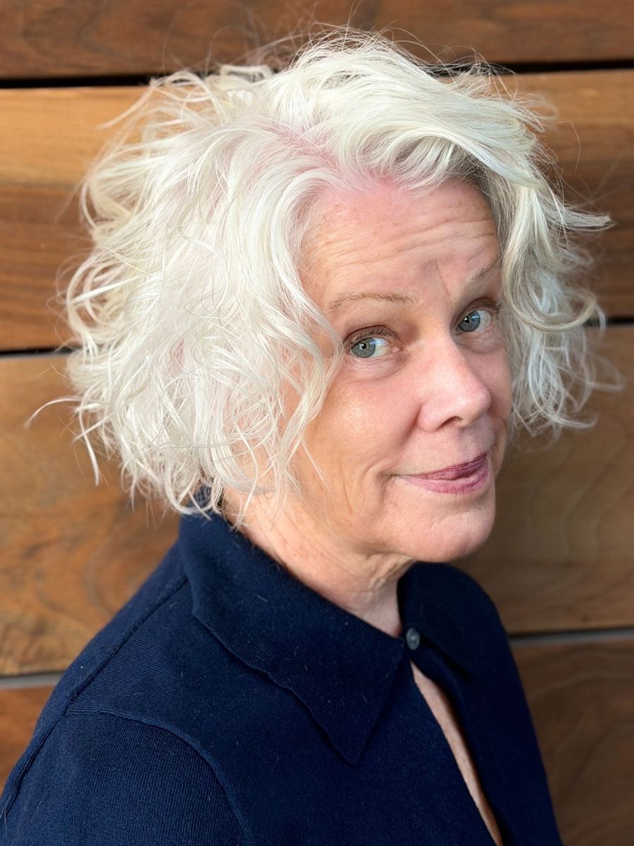 Close-up portrait of an older woman with short, curly white hair and blue eyes, wearing a navy collared shirt, in front of a wooden backdrop.