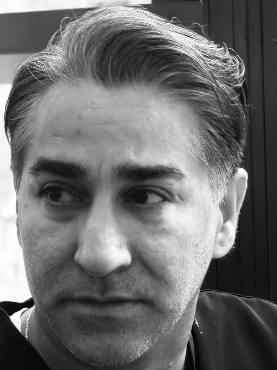 Close-up of a man with short, slicked-back hair and light stubble, looking to his left against a dark background.