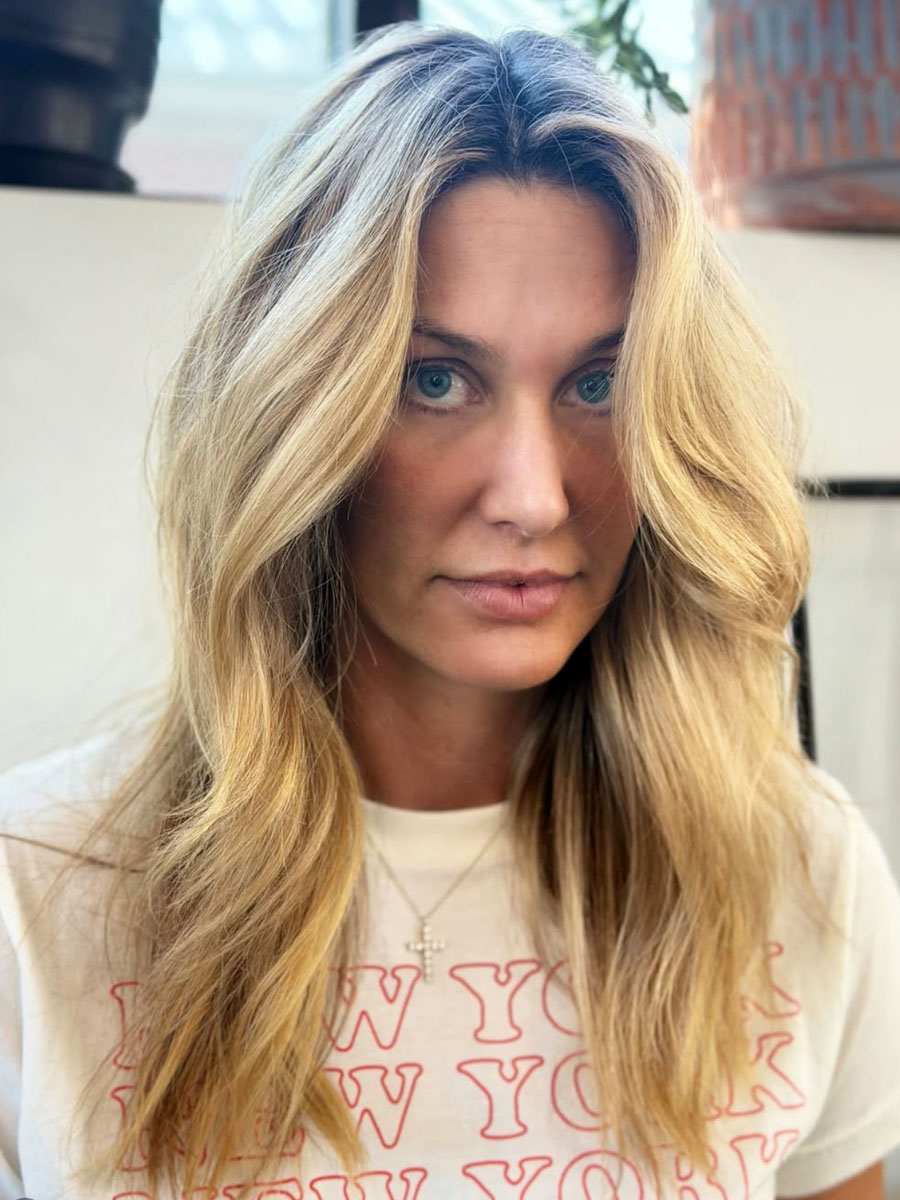 Close-up of a woman with long blonde hair and blue eyes, wearing a white t-shirt with red text and a cross necklace outdoors.