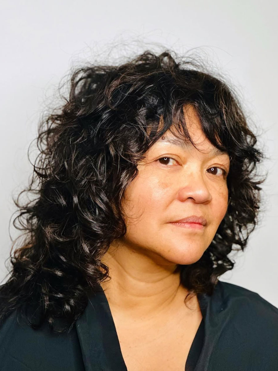 Portrait of a middle-aged woman with dark, curly hair wearing a dark top, facing slightly to the right with a neutral expression on a light background.